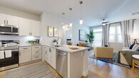 This well-designed single-floor residence offers a bright, open-concept layout that seamlessly connects the kitchen, dining, and living areas. The kitchen is a standout feature, equipped with white shaker-style cabinetry, granite countertops, stainless steel appliances including a side-by-side refrigerator, range, and built-in microwave, and a central island with pendant lighting overhead and a stainless steel sink. Light wood-style flooring runs throughout the main living areas, enhancing the airy, contemporary feel. The living room is generously sized, filled with natural light from large windows with plantation shutters, and features a ceiling fan for added comfort. Multiple bedrooms are present throughout the residence, each carpeted and outfitted with ceiling fans and windows that bring in ample natural light. The primary bedroom includes access to a private en-suite bathroom featuring a walk-in shower with tile surround, a granite-topped vanity with white cabinetry, and wood-style flooring. A secondary bathroom mirrors the refined finishes with a granite vanity, tub-shower combination, and tile surround. The overall ambiance throughout is clean, modern, and cohesive, with a neutral color palette that creates a welcoming and functional living environment.