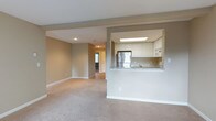 This single-floor residence offers a thoughtfully designed open-concept layout that seamlessly connects the living area and kitchen. The living space is generously sized, finished with soft neutral-toned carpeting and warm beige walls that create a comfortable, welcoming ambiance. Recessed lighting is incorporated into the ceiling, complementing the natural light that filters through the space.

The kitchen is positioned behind a half-wall pass-through, which features a light stone or laminate countertop that doubles as a convenient breakfast bar. White cabinetry lines the upper walls of the kitchen, providing ample storage, while a stainless steel refrigerator serves as a standout appliance. A skylight above the kitchen floods the area with additional natural light, making it feel bright and airy. Built-in microwave and additional appliances are neatly integrated within the cabinetry. Tile backsplash adds a clean, finished detail to the kitchen walls.

A hallway visible from the main living area leads to additional rooms, suggesting a practical and well-connected floor plan. The clean white trim and baseboards throughout provide a crisp contrast to the neutral wall tones, contributing to an overall aesthetic that is both functional and inviting.