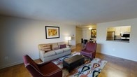 This single-floor space presents a thoughtfully arranged living area that serves as the central hub of the home. The living room is furnished with a beige upholstered sofa accented by decorative throw pillows in warm red and neutral tones, complemented by a pair of deep burgundy accent chairs with white piping detail. A large rectangular black leather ottoman anchors the seating arrangement atop a colorful geometric-patterned area rug in shades of blue, red, and gold, adding visual interest to the hardwood flooring beneath. A framed piece of artwork in warm amber and ochre tones hangs prominently on one wall, contributing to the cohesive warm color palette throughout. A classic table lamp provides soft ambient lighting in the corner. The space opens seamlessly into a dining area, visible in the background, where a ceiling fan with integrated light fixture illuminates a modest dining table with dark wood chairs. A second framed artwork echoes the living room's aesthetic in the dining space. The overall ambiance is warm, inviting, and well-coordinated, with a layout that maximizes the sense of openness across the single floor.