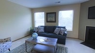 This single-floor residence offers a clean, comfortable, and well-appointed living environment designed with everyday functionality in mind. The living room serves as the heart of the space, bathed in natural light streaming through multiple windows fitted with blinds for adjustable privacy. Neutral beige walls provide a versatile backdrop that complements the deep navy sectional sofa, accented with patterned throw pillows that add a touch of personality. A sturdy rectangular coffee table with an industrial-style metal frame anchors the seating area, resting atop a textured area rug that adds warmth to the light-toned flooring beneath. Decorative touches, including a framed nature-inspired artwork centered between the windows and a globe accent piece on the table, lend the room a curated yet relaxed aesthetic. A potted plant in the corner introduces a subtle natural element, enhancing the overall sense of calm. The space benefits from a simple, uncluttered layout that maximizes openness and ease of movement. Overall, this residence strikes a balance between practical living and inviting comfort, making it an ideal setting for both relaxing and entertaining.