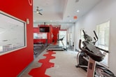 Located at 105 Old Hull Rd in Athens, Georgia, this fitness center is a well-appointed exercise facility designed to support a full range of workout routines. The space is divided into two distinct zones, each with its own character and function. The main cardio area features crisp white walls, recessed lighting, and large floor-to-ceiling mirrors that amplify the sense of space. A bold, geometric red-and-gray patterned carpet runs through the center of the floor, adding a dynamic visual element. Cardio equipment, including treadmills and an elliptical machine, is thoughtfully arranged to allow comfortable use, while multiple windows and a glass door invite natural light and views of the surrounding greenery outside. A dedicated strength and free weights area provides dumbbell racks in varying weights, medicine balls, stability balls, and a weight bench, catering to a variety of strength training needs. The second zone features striking bold red walls, a ceiling fan, and pass-through windows that connect visually to the main area, creating an energetic and motivating atmosphere ideal for high-intensity workouts. A wall-mounted television and designated free-weight space round out this area. Together, both zones offer a comprehensive, well-equipped fitness environment.