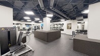 Located at 1400 N Lake Shore Drive in Chicago's Gold Coast neighborhood, this well-appointed fitness center offers a comprehensive workout facility across a single floor. The space is defined by its industrial-modern aesthetic, featuring a striking black drop-tile ceiling with exposed ductwork and ample recessed lighting that keeps the environment bright and energizing.

The main workout area is expansive and open, with durable speckled rubber flooring ideal for heavy equipment use. It houses a wide selection of cardio machines — including Life Fitness treadmills, ellipticals, and stationary bikes — as well as a robust lineup of selectorized strength machines and a dedicated free weights section with a full dumbbell rack. Floor-to-ceiling mirrors line the walls, amplifying the sense of space and supporting proper form during exercise. Wall-mounted TVs are positioned throughout for entertainment during workouts.

A separate studio room features elegant herringbone hardwood flooring and mirrored walls, making it well suited for group fitness classes, stretching, or personal training sessions. This area is also stocked with functional training accessories including stability balls, kettlebells, resistance bands, and a multi-functional rack.

Throughout the facility, white structural columns punctuate the layout, and framed artwork adds a refined touch to the utilitarian setting. The overall design balances functionality with a polished, professional atmosphere.