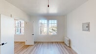 This single-floor unit presents a clean, modern aesthetic anchored by light hardwood flooring that runs throughout the living areas. Crisp white walls and multiple gridded windows create a bright, airy atmosphere, allowing natural light to flow freely across the space.

The living area is generously sized, featuring a ceiling fan and a classic cast-iron radiator beneath a large street-facing window that frames urban views of the surrounding neighborhood. A pull-cord pendant light adds a subtle vintage touch to the otherwise minimalist room.

The kitchen is efficiently designed with navy blue cabinetry, white countertops, a stainless steel sink, a gas range with an overhead exhaust hood, and a white subway tile backsplash. A pass-through opening connects the kitchen to the adjoining living space, enhancing the open feel. A ceiling fan and dedicated window keep the kitchen well-ventilated and naturally lit.

The bathroom showcases floor-to-ceiling white subway tile with dark grout lining the tub surround, complemented by matte black fixtures including the faucet, tub spout, and showerhead. A window above the tub invites natural light while maintaining a sleek, contemporary look.

Overall, the unit offers a cohesive, refreshed interior with thoughtful details and a bright, welcoming ambiance throughout.