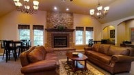 This single-floor space exudes a warm, traditional charm from the moment you step inside. The living area serves as the heart of the home, anchored by a commanding floor-to-ceiling stone fireplace with a classic brick hearth and a rich wood mantel — an undeniable focal point that lends rustic elegance to the room. Vaulted ceilings rise overhead, enhancing the sense of openness and grandeur, while recessed lighting and two decorative chandelier-style fixtures cast a welcoming glow throughout the space. Generous windows with dark valances frame views of lush greenery outside, flooding the interior with natural light and connecting the indoors to the surrounding landscape. The living area is furnished with plush leather sofas arranged around a traditional wood coffee table, all set atop a detailed area rug that adds texture and warmth to the space. Warm golden tones on the walls complement the earthy stone and wood accents, creating a cohesive and inviting color palette. A dining area is visible in the background, hinting at a seamlessly flowing layout ideal for both everyday living and entertaining.