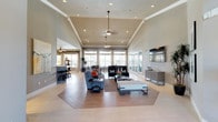 This single-floor space is designed to impress with its grand, open-concept layout and refined architectural details. The main living and lounge area commands attention with soaring vaulted ceilings adorned with crisp crown molding and a raised tray ceiling section, creating a sense of grandeur and volume. Dual ceiling fans and recessed lighting throughout ensure the space remains both bright and comfortable at any time of day.

The flooring is a standout feature, showcasing a striking herringbone-patterned tile inset set against large-format light-toned tile, adding a sophisticated geometric focal point to the room. Floor-to-ceiling windows along the far wall flood the interior with abundant natural light, while sheer drapery provides a soft, elegant finish.

The lounge area is furnished with plush seating arrangements, complemented by a media console and wall-mounted flat-screen television. Adjacent to the main lounge, a stylish bar or café area with counter seating offers an inviting secondary gathering space, visible through an open archway.

The overall ambiance is modern yet welcoming, blending clean architectural lines with warm, neutral tones. This well-appointed space is ideal for both relaxed gatherings and more structured social events.