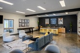 Located at 901 S Harbor Blvd in Santa Ana, California, this single-floor community space is designed with a contemporary aesthetic that blends functionality and social appeal. The open-plan layout is anchored by polished concrete floors, exposed dark ceiling beams, and a striking contrast between crisp white walls and bold charcoal accent walls that lend the space a modern, energetic character.

The lounge area serves as the centerpiece, furnished with plush modular seating, rounded accent chairs, and woven area rugs arranged around a low-profile round coffee table. A wall-mounted flat-screen TV enhances the entertainment zone, while a pair of rattan hanging chairs positioned against the dark accent wall add a relaxed, bohemian touch. Natural light filters through multiple sliding windows and glass-paned doors that open to outdoor views.

A dedicated kitchenette runs along one wall, featuring dark granite countertops, warm wood cabinetry, a double basin sink, and bar-height seating. A glowing neon "Good Vibes Only" sign mounted above the counter adds personality and sets an upbeat tone throughout the space. Additional amenities include a shuffleboard table and gallery-style wall art, making this a well-rounded hub for socializing and recreation. Recessed and flush panel lighting ensure the space is bright and welcoming at all hours.