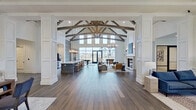 This impressive single-floor amenity space combines elegant architectural design with versatile, open-concept functionality. The centerpiece of the space is a dramatic vaulted ceiling adorned with rustic exposed wood beam trusses, soaring high above wide-plank hardwood floors that run throughout. Towering floor-to-ceiling windows and black-framed glass doors flood the interior with natural light while offering views of the surrounding community.

The open main hall flows seamlessly into a well-appointed kitchen featuring a large island with bar seating and pendant lighting, positioned alongside a spacious great room anchored by a classic fireplace and comfortable lounge seating. Decorative wainscoting, white panel millwork, and structural columns add refined architectural detail throughout.

A dedicated office or reception area includes coffered ceilings, a globe chandelier, large mirrors, and an executive desk setup, creating a professional yet welcoming environment. Adjacent lounge areas offer additional flexible seating arrangements beneath elegant chandeliers and coffered ceiling details.

The game room provides entertainment with a foosball table, billiards, and built-in shelving flanking a fireplace, with barn-style doors leading to an outdoor patio. The overall ambiance is sophisticated and inviting, blending modern farmhouse aesthetics with upscale community living.