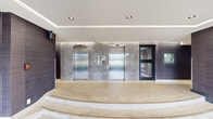 Located at 2150 Roche Crt in Mississauga, Ontario, this building lobby presents a refined and contemporary aesthetic that sets a welcoming tone for residents and visitors alike. The space is defined by its polished terrazzo flooring, which features warm neutral tones with subtle aggregate detailing, extending throughout the entire lobby area and creating a seamless, elegant foundation underfoot.

The elevator bank serves as a focal point within the lobby, featuring two stainless steel elevator doors set against a backdrop of large-format marble-effect wall tiles in soft grey tones, lending the space a sophisticated, upscale character. Gently curved terrazzo steps lead up to the elevator landing, adding architectural interest and visual depth to the layout.

Contrasting wall treatments — including richly textured dark wood-toned panels — provide warmth and dimension throughout the space. A dedicated mailroom area is neatly integrated into the lobby, featuring an extensive bank of stainless steel mailboxes mounted against a striking dark fluted wall panel, combining functionality with modern design.

Floor-to-ceiling glazed entrance doors and sidelights flood the lobby with natural light and offer open views of the surrounding landscaped grounds. Recessed ceiling lighting, complemented by a clean coffered ceiling detail with subtle cove illumination, ensures the space is well-lit and polished at all times. Overall, the lobby exudes a calm, professional ambiance suited to a well-maintained residential building.