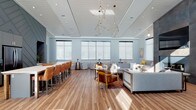 This single-floor amenity space is thoughtfully designed to combine comfort, functionality, and contemporary style throughout its open-concept layout. The main lounge area immediately impresses with soaring vaulted ceilings clad in white shiplap, dramatic geometric pendant light fixtures in brass-toned metal, and rich hardwood flooring that flows throughout the gathering spaces. Expansive clerestory windows and large roller-shade windows flood the interior with abundant natural light, creating a bright and welcoming atmosphere.

The main lounge features multiple seating arrangements, including plush gray sofas, tufted accent chairs, and leather bar-height seating along a full kitchen bar. The kitchen area is appointed with stainless steel appliances, cabinetry in a deep slate finish, and a chevron-patterned accent wall that serves as a bold architectural focal point. An adjoining lounge area offers a more intimate setting, centered around a floor-to-ceiling tile-clad fireplace with a built-in wall-mounted television and a cozy built-in window bench with accent cushions.

A separate office suite provides a professional environment with plush carpeting, dark-toned walls, workstation desks, and a television display area, offering a quiet and focused space distinct from the social areas. Throughout, the design strikes a balance between modern sophistication and approachable warmth.
