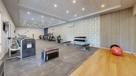 Located at 55 Tunxis Ave in Bloomfield, Connecticut, this fitness center offers a generous, open single-floor layout designed to accommodate a full range of exercise activities. The space is thoughtfully organized to maximize functionality and flow, with distinct zones dedicated to cardio training, strength work, and functional fitness.

The cardio section features a row of treadmills and elliptical cross-trainers from brands such as Matrix, providing ample options for endurance-focused workouts. Adjacent to this area, a strength training zone is equipped with a dumbbell rack, weight benches, and resistance machines, catering to users of all fitness levels. A dedicated functional training area includes plyo boxes, medicine balls, and open floor space with hardwood flooring — ideal for dynamic movement and group exercise.

The interior design is modern and polished, with a drop tile ceiling outfitted with recessed lighting that creates a bright, energizing atmosphere throughout. A distinctive geometric-patterned accent wall adds visual interest, while large windows along one side invite natural light and a sense of openness. Wall-mounted televisions and a wall-to-wall mirror further enhance the environment. The durable, rubber-composite flooring throughout the main workout area ensures safety and comfort during training sessions.