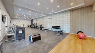 Located at 55 Tunxis Ave in Bloomfield, Connecticut, this fitness center offers a generous, open single-floor layout designed to accommodate a full range of exercise activities. The space is thoughtfully organized to maximize functionality and flow, with distinct zones dedicated to cardio training, strength work, and functional fitness.

The cardio section features a row of treadmills and elliptical cross-trainers from brands such as Matrix, providing ample options for endurance-focused workouts. Adjacent to this area, a strength training zone is equipped with a dumbbell rack, weight benches, and resistance machines, catering to users of all fitness levels. A dedicated functional training area includes plyo boxes, medicine balls, and open floor space with hardwood flooring — ideal for dynamic movement and group exercise.

The interior design is modern and polished, with a drop tile ceiling outfitted with recessed lighting that creates a bright, energizing atmosphere throughout. A distinctive geometric-patterned accent wall adds visual interest, while large windows along one side invite natural light and a sense of openness. Wall-mounted televisions and a wall-to-wall mirror further enhance the environment. The durable, rubber-composite flooring throughout the main workout area ensures safety and comfort during training sessions.