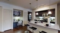 This single-floor residence is designed with a contemporary aesthetic that blends functionality and style throughout. The heart of the home is an open-concept kitchen that flows seamlessly into the living areas, creating an inviting and connected atmosphere. The kitchen is outfitted with rich, dark cabinetry that contrasts elegantly against light granite countertops, which extend along a generous breakfast bar complete with sleek black bar stools. Stainless steel appliances — including a refrigerator, range, and microwave — lend a polished, modern character to the space. Pendant lighting hangs above the kitchen island, while track lighting along the ceiling provides additional illumination, ensuring the space is both well-lit and visually dynamic. The floors throughout are finished in dark hardwood, adding warmth and sophistication underfoot. Crown molding along the ceiling contributes a refined architectural detail. A thoughtfully designed built-in workstation nook sits just off the kitchen, featuring dedicated cabinetry, under-cabinet lighting, and space for a desk — perfect as a home office or study area. Decorative wall art adds a personalized touch to this alcove. The overall ambiance is modern and well-appointed, offering a cohesive and comfortable living environment.