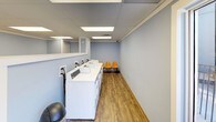 Mountainside Laundry is a single-floor commercial laundry facility designed for practicality and comfort. The space is finished with soft blue-painted walls and durable wood-look flooring throughout, creating a clean and welcoming environment. Overhead, a drop ceiling with large flush-mount panel lights ensures the room is evenly and brightly illuminated.

A row of commercial-grade top-load washing machines lines one wall, positioned beneath a mirrored upper section that adds a sense of openness to the space. Adjacent to the washers, a set of stacked commercial dryers — each individually labeled and equipped with digital control panels — provides ample drying capacity. A bold yellow folding table with metal hairpin legs offers a dedicated surface for sorting and folding laundry, while a set of molded orange chairs along the opposite wall provides a comfortable waiting area for patrons.

The layout is straightforward and efficiently organized, with clear circulation space between the equipment and seating. A waste bin is conveniently positioned near the entrance, and ventilation is handled through ceiling and wall registers. The overall ambiance is tidy and functional, reflecting a well-maintained facility suited to meet the everyday laundry needs of its users.