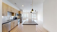 This single-floor residence offers a thoughtfully designed open-concept layout that seamlessly connects the kitchen, dining, and living areas. The kitchen is a standout feature, outfitted with light wood cabinetry, dark granite countertops, a herringbone-patterned tile backsplash, and a full suite of stainless steel appliances including a refrigerator, electric range, microwave, and dishwasher. A spacious center island with a dark stone countertop serves as a natural focal point, accented by sleek pendant lighting overhead. Light wood flooring runs throughout the main living areas, complementing the bright, white walls and recessed lighting that give the space an airy, modern feel. The open living area benefits from multiple windows that invite natural light, and is equipped with a ceiling fan for added comfort. The bedrooms feature plush carpeting, vaulted ceilings, and generous windows. The primary bedroom connects directly to an en-suite bathroom, which includes a wood-toned vanity with granite countertops, a soaking tub with marble-style tile surround, and a walk-in closet. A dedicated in-unit laundry area with a full washer and dryer adds everyday convenience. The cohesive design and quality finishes throughout make this a well-appointed and functional living space.