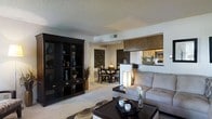 This single-floor space showcases an open-concept design that seamlessly connects the living, dining, and kitchen areas into one cohesive environment. The living area is anchored by a plush sectional sofa in a soft neutral tone, paired with a sleek dark-wood coffee table that adds contrast and visual weight. A large, dark display shelving unit lines one wall, offering both storage and decorative space, and is styled with vases, greenery, and accent pieces that contribute to the room's refined aesthetic.

Adjacent to the living area, the dining space features a round dark-wood table surrounded by upholstered chairs, creating an intimate setting for meals. Decorative wall art with geometric patterns adds depth and character to the space. A floor lamp with a warm-toned shade provides soft accent lighting throughout.

A half-wall partition separates the kitchen from the main living area, offering a pass-through view while maintaining a sense of openness. The kitchen features warm cabinetry and appears well-appointed. Throughout the space, carpeted flooring, a textured ceiling, and warm lighting combine to create a comfortable and contemporary ambiance.