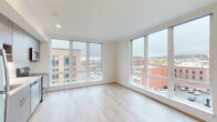 This modern urban unit is thoughtfully designed with a clean, contemporary aesthetic and an abundance of natural light. The open living area is defined by large, floor-to-ceiling windows positioned on two walls, framing expansive views of the surrounding cityscape, including neighboring mixed-use buildings and street-level activity below. Light wood-tone flooring runs throughout the unit, contributing to a bright and cohesive atmosphere.

The kitchen is efficiently laid out along a single wall and features flat-front cabinetry in a warm wood-grain finish, light-colored quartz countertops, a stainless steel sink, and a full suite of stainless steel appliances including a range, over-the-range microwave, and dishwasher. A large window adjacent to the kitchen provides additional natural light and urban views.

The bathroom offers a clean, modern finish with a wood-toned vanity, white quartz countertop, a large mirror, and a combination tub and shower. Wood-look flooring continues into this space for a consistent design language throughout. A dedicated in-unit laundry closet houses a stacked washer and dryer. The entry corridor features warm-toned hardwood flooring and a charcoal-finished door, offering a welcoming transition into the unit.