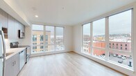 This modern urban unit is thoughtfully designed with a clean, contemporary aesthetic and an abundance of natural light. The open living area is defined by large, floor-to-ceiling windows positioned on two walls, framing expansive views of the surrounding cityscape, including neighboring mixed-use buildings and street-level activity below. Light wood-tone flooring runs throughout the unit, contributing to a bright and cohesive atmosphere.

The kitchen is efficiently laid out along a single wall and features flat-front cabinetry in a warm wood-grain finish, light-colored quartz countertops, a stainless steel sink, and a full suite of stainless steel appliances including a range, over-the-range microwave, and dishwasher. A large window adjacent to the kitchen provides additional natural light and urban views.

The bathroom offers a clean, modern finish with a wood-toned vanity, white quartz countertop, a large mirror, and a combination tub and shower. Wood-look flooring continues into this space for a consistent design language throughout. A dedicated in-unit laundry closet houses a stacked washer and dryer. The entry corridor features warm-toned hardwood flooring and a charcoal-finished door, offering a welcoming transition into the unit.