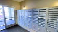 The mail room is a functional, single-floor common area designed to serve the building's residents efficiently. A full wall is lined with rows of individual metal mailboxes finished in a light gray tone, each equipped with its own keyed lock for secure, private mail access. The units are organized in a grid arrangement, accommodating a large number of residents. Several larger parcel compartments are integrated at the base of the unit clusters, providing additional storage for package deliveries. The room features neutral beige walls and carpeted flooring in a muted, speckled pattern that contributes to a clean, practical aesthetic. A wall-mounted light fixture provides warm, ambient illumination. Natural light filters in from a window near the entrance, brightening the space. The overall design prioritizes utility and organization, offering residents a straightforward and accessible location to retrieve their mail and packages within the building.