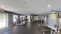 This single-floor fitness center is designed to support a full range of workout routines in a spacious, open layout. The interior is finished with cool blue-gray walls, a textured ceiling with recessed lighting throughout, and durable dark rubber flooring that defines the exercise areas. Large windows fitted with white blinds bring in generous natural light, while a set of glass double doors opens to views of an outdoor pool, creating an airy, connected feel.

The cardio section is well-stocked with treadmills, elliptical trainers, a stair climber, and Peloton-style stationary bikes. A large wall mirror reflects the space, enhancing the sense of openness. For strength training, the facility offers a multi-function cable and pull-up station, an adjustable weight bench, and a full dumbbell rack with wall-mounted televisions nearby. A separate, quieter alcove area provides additional floor space for stretching or functional training, decorated with framed artwork and equipped with an ab bench. The room also includes safety features such as a fire extinguisher and emergency exit signage. Overall, the fitness center delivers a clean, functional, and motivating environment for daily exercise.