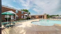 This outdoor pool facility in Marlborough, Missouri offers a well-appointed recreational retreat designed for private use. At its centerpiece is a large, freeform swimming pool with smooth curvilinear edges and clear, inviting water, framed by a broad concrete sundeck that provides plenty of open space for movement and relaxation. Rows of green lounge chairs are arranged throughout the deck, offering abundant seating for sunbathing and leisure. The pool area is fully enclosed by a black metal fence with a secured gate entry, ensuring privacy and controlled access. A posted pool regulations sign at the entrance outlines rules and hours of operation. Mature trees and neatly maintained landscaping with mulched garden beds border portions of the perimeter, adding greenery and a sense of seclusion. The surrounding multi-story brick residential buildings frame the space on multiple sides, contributing to a cohesive, community-oriented atmosphere. Open skies above the deck allow for ample natural light throughout the day, enhancing the overall outdoor ambiance. The combination of a spacious pool, well-organized seating, and thoughtfully maintained grounds makes this a functional and attractive amenity for residents of the community.