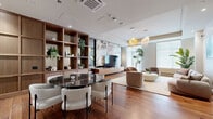 Located at 3000 Rue Modugno in Montréal, this elegantly designed lounge offers a refined open-concept layout that seamlessly blends comfort and contemporary sophistication. Rich hardwood flooring flows throughout the entire space, anchoring a warm, inviting atmosphere complemented by crisp white ceilings with recessed and track lighting.

A striking floor-to-ceiling marble accent wall serves as the visual centerpiece, framing a wall-mounted television and sleek media console. Adjacent to it, a full-height walnut shelving unit provides both display and storage, adding warmth and structure to the open plan. The dining area features a round dark marble-topped table surrounded by curved white upholstered chairs, positioned conveniently near a kitchenette with light cabinetry and brass fixtures.

The lounge seating area is furnished with a generous curved sofa in neutral tones, sculptural rounded armchairs in warm brown, and an organic-shaped white coffee table, all arranged atop a textured area rug. Oversized windows dressed with sheer roller shades bathe the space in diffused natural light, while tall indoor plants add a refreshing natural element throughout. Pendant lighting with woven gold shades adds an artful finishing touch, completing this polished and functional lounge environment.