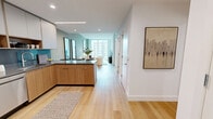 Located at 5 Cottage Place in White Plains, NY, this thoughtfully designed one-bedroom, one-bathroom residence offers a sleek, modern aesthetic across a single floor. The open-concept layout seamlessly connects the kitchen, dining, and living areas, all unified by warm light wood flooring and a soft, neutral palette. The contemporary kitchen features two-tone cabinetry — combining white upper cabinets with natural wood lower cabinets — dark stone countertops, a teal backsplash, stainless steel appliances, and an island that doubles as a dining peninsula. The living room is anchored by expansive floor-to-ceiling windows that flood the space with natural light and offer sweeping views of the White Plains skyline. The bedroom continues the window-forward design with corner glass panels framing panoramic city views, creating a bright and airy retreat. A versatile den provides additional flex space, ideal as a home office. The bathroom is finished with large-format gray tile, a wood-framed vanity with stone countertop, a lit mirror, and a glass-enclosed walk-in shower. Two well-organized closets with adjustable shelving systems and a dedicated laundry area complete this well-appointed residence.