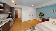 Located at 259 Ferncliff Ave NE on Bainbridge Island, WA, this single-floor studio offers a well-designed, efficient layout that makes excellent use of its space. The interior is finished with warm hardwood-style flooring throughout, clean white walls, and recessed lighting complemented by track lighting near the entryway, creating a bright and welcoming atmosphere.

The main living and sleeping area is anchored by a distinctive teal accent wall, adding a pop of color to the otherwise neutral palette. Large windows dressed with white horizontal blinds allow generous natural light to fill the space. A cozy seating area features a plush sofa with decorative pillows and a wall-mounted flat-screen television, while a small dining nook with vibrant yellow chairs provides a functional yet stylish corner for meals.

The kitchen is outfitted with dark, textured cabinetry, light quartz countertops, a stainless steel undermount sink with a flexible faucet, and a tile backsplash. Open shelving above the counters provides accessible storage, and stainless steel appliances complete the contemporary culinary space.

Additional features include multiple closets for ample storage, a private entry door, and an overall mid-century modern aesthetic that ties the space together cohesively. The layout is open and versatile, ideal for comfortable single-occupancy living on Bainbridge Island.
