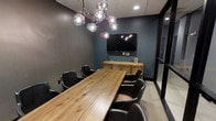 This single-floor professional space is designed to impress, combining modern aesthetics with functional design. The centerpiece of the space is a sophisticated conference room that exudes a polished, contemporary atmosphere. Deep blue-gray accent walls paired with textured wallcoverings create a rich, focused environment ideal for meetings and collaborative sessions. A large, solid wood conference table serves as the anchor of the room, surrounded by sleek black leather chairs that offer both comfort and a professional look. A wall-mounted flat-screen display positioned above a warm wood credenza provides seamless technology integration for presentations and video conferencing. The room is crowned by a striking multi-globe chandelier with a sculptural, branching design that adds an artistic focal point and a touch of elegance to the space. Recessed lighting complements the chandelier to ensure the room is well-lit throughout. Glass partition elements along the perimeter allow for visual connectivity while maintaining privacy, contributing to a sense of openness within the overall layout. The combination of natural wood tones, dark upholstery, and moody wall colors gives this space a refined, executive character suited for professional use.
