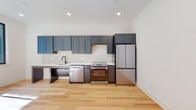 This single-floor unit showcases a thoughtfully designed, contemporary interior with consistent hardwood flooring in a warm, light natural finish running throughout the space. The kitchen serves as a central feature, outfitted with floor-to-ceiling slate-blue and grey shaker-style cabinetry offering generous storage. A clean white countertop extends along the full length of the wall, complemented by a subway tile backsplash and a matte black faucet at the undermount sink. Stainless steel appliances include a dishwasher, range, and a full-size French door refrigerator. Recessed lighting illuminates the kitchen from above, contributing to a bright, open atmosphere.

The bathroom presents a modern aesthetic with large hexagonal grey floor tiles and white subway wall tiles extending partway up the walls. A wall-mounted sink pairs with a distinctive arched black metal mirror, and matte black fixtures and grab bars add both style and accessibility. A glass-enclosed shower is also present.

The bedroom is bright and minimalist, with white walls, hardwood floors, and a black-framed window that draws in natural light. The overall layout, as seen in the 3D overview, is open and efficiently arranged, creating a comfortable and functional living environment.