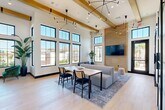Located at 7631 Calloway Dr in Bakersfield, CA, this single-floor community space offers a thoughtfully designed interior that blends modern farmhouse aesthetics with contemporary finishes. The main living area immediately impresses with dramatically high ceilings adorned with exposed natural wood beams and striking sculptural chandelier lighting. A wall of floor-to-ceiling black-framed windows floods the space with abundant natural light while framing views of the surrounding outdoor amenities, including palm trees and a pool area. Light hardwood flooring runs throughout, lending warmth and continuity to the open layout. A full-height wood-paneled accent wall anchors the lounge area, where a modular gray sofa and mixed seating arrangements are centered around a natural wood dining table. A marble-topped kitchen island with pendant lighting defines a dedicated refreshment area, adding an upscale touch. Sliding barn doors with frosted glass panels connect to a private nook, where bold teal and geometric patterned wallcoverings create an intimate, design-forward retreat. Lush potted plants are strategically placed throughout, contributing to a fresh, inviting ambiance. This versatile, well-appointed space is ideal for gathering, socializing, or focused work.