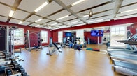 This fitness center is a comprehensively outfitted workout facility designed to support a wide range of exercise routines. The space is immediately striking thanks to its bold deep-red walls, which create an energetic and motivating atmosphere throughout. Light-toned hardwood-style flooring runs across the entire floor, providing a clean, durable surface suitable for both equipment use and open-floor training activities.

The ceiling features a distinctive coffered design with exposed beams and integrated fluorescent lighting, contributing to a bright and well-illuminated environment. Ceiling fans are also present, aiding in air circulation during workouts. Multiple windows allow natural light to filter in, further enhancing the open feel of the space.

The facility is divided into functional workout zones. A dedicated cardio area houses multiple treadmills and upright cycling machines, each equipped with individual display screens. The strength training section includes a cable crossover machine, a multi-functional rack system, an adjustable bench, and various resistance machines. A free weights station offers a full rack of dumbbells alongside a water station for convenience.

A floor-to-ceiling mirror spans one wall, visually expanding the space and allowing users to monitor their form. Functional training equipment — including kettlebells, medicine balls, resistance bands, and foam rollers — is neatly organized throughout. Multiple flat-screen televisions are mounted for entertainment during workouts, completing this well-rounded fitness facility.