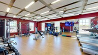 This fitness center is a comprehensively outfitted workout facility designed to support a wide range of exercise routines. The space is immediately striking thanks to its bold deep-red walls, which create an energetic and motivating atmosphere throughout. Light-toned hardwood-style flooring runs across the entire floor, providing a clean, durable surface suitable for both equipment use and open-floor training activities.

The ceiling features a distinctive coffered design with exposed beams and integrated fluorescent lighting, contributing to a bright and well-illuminated environment. Ceiling fans are also present, aiding in air circulation during workouts. Multiple windows allow natural light to filter in, further enhancing the open feel of the space.

The facility is divided into functional workout zones. A dedicated cardio area houses multiple treadmills and upright cycling machines, each equipped with individual display screens. The strength training section includes a cable crossover machine, a multi-functional rack system, an adjustable bench, and various resistance machines. A free weights station offers a full rack of dumbbells alongside a water station for convenience.

A floor-to-ceiling mirror spans one wall, visually expanding the space and allowing users to monitor their form. Functional training equipment — including kettlebells, medicine balls, resistance bands, and foam rollers — is neatly organized throughout. Multiple flat-screen televisions are mounted for entertainment during workouts, completing this well-rounded fitness facility.