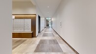 Located at 825 Fort St in the heart of Victoria, BC, this ground-floor lobby area comprises two interconnected hallways that together form a welcoming and well-appointed entry experience. The L-shaped layout, as revealed by the dollhouse view, connects the building's main entrance to its elevator and stairwell access points with a clean, purposeful flow.

The primary hallway is distinguished by its striking herringbone-patterned hardwood flooring, flanked by charcoal-grey wainscoting with crisp panel detailing and warm natural wood millwork. Elegant pendant light fixtures hang from the elevated white ceiling, casting a refined glow throughout. An integrated bank of individual mailboxes, set within a rich wood-framed surround with warm accent lighting, adds a functional yet stylish focal point. Open shelving adds further practical character to this corridor.

The secondary hallway transitions to large-format porcelain tiles laid in a checkerboard-style pattern of varying neutral tones, maintaining a bright and airy atmosphere with clean white walls and dark wood baseboards. Glass-panel doors at the entrance allow natural light to penetrate deep into the space, enhancing the sense of openness. Recessed ceiling lights and a clear sightline toward the building exterior complete the polished, contemporary aesthetic of this well-designed communal entryway.