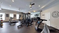 Located in Jefferson, this single-floor fitness center is designed to support a comprehensive workout experience. The space is outfitted with a robust selection of equipment, including a multi-station cable and weight machine, cardio machines such as treadmills and ellipticals, and flat benches for free weight exercises. The rubber flooring throughout provides both safety and durability, making it well-suited for high-intensity training. The ceiling is fitted with recessed lighting and multiple ceiling fans, ensuring the space stays bright and well-ventilated during workouts. A large wall-to-wall mirror along one side of the room visually expands the area and allows users to monitor their form. Natural light filters in through windows on the far wall, contributing to an energizing atmosphere. The neutral gray walls and dark equipment create a focused, functional aesthetic throughout. The layout is thoughtfully organized to allow for comfortable movement between stations, making this fitness center a practical and fully equipped facility for residents or members.