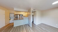This single-floor unit offers a thoughtfully designed open-concept layout that seamlessly connects the kitchen and living areas. The kitchen is outfitted with white shaker-style cabinetry, stainless steel appliances including a refrigerator, built-in microwave, and dishwasher, complemented by a classic white subway tile backsplash and a granite-style countertop that extends into a breakfast bar. A stacked washer and dryer unit is conveniently integrated within the kitchen space, adding practical in-unit laundry access. Light wood-tone flooring runs throughout the main living areas, contributing to a clean and contemporary aesthetic. The living area is generously proportioned, featuring a ceiling fan with integrated lighting and a sliding glass door that opens onto a private balcony, allowing natural light to fill the space and offering views of surrounding greenery. The bathroom is finished with white tile surrounding a full tub-and-shower combination, wood-look flooring, and clean, minimal fixtures. A walk-in closet is accessible from the main living area, providing ample storage. The overall ambiance is bright, neutral, and move-in ready, with a functional and efficient layout suited for everyday living.