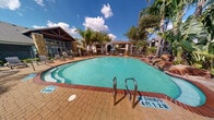 This outdoor pool reserve is a well-appointed amenity space designed for relaxation and recreation. At its center sits a large, freeform swimming pool with clear turquoise water, featuring gradual entry points, stainless steel handrails, and clearly marked depth indicators. The pool's organic shape creates an inviting, resort-like atmosphere that encourages leisurely use throughout the day.

Surrounding the pool is a spacious brick-tiled deck generously outfitted with lounge chairs, providing ample space for sunbathing and outdoor leisure. Shaded seating areas with patio furniture and market umbrellas offer comfortable alternatives for those seeking relief from direct sunlight.

The landscaping throughout the reserve is lush and tropical in character, with tall palm trees, mature shade trees, and decorative rock formations adding visual depth and a resort-style ambiance. Natural light floods the space throughout the day, enhanced by the open-sky setting.

Adjacent to the pool deck stands a clubhouse structure with a distinctive gabled roofline, exposed timber beams, and stone-accented exterior columns, complemented by expansive windows that connect the interior to the outdoor environment. A pergola structure and additional seating areas are also present, further enhancing the usability of the space. The entire area is enclosed by a perimeter fence, ensuring a secure and private environment for residents.