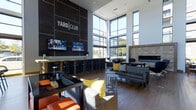 The Yard Club is a contemporary communal lounge space located at 845 Yard St in Grandview Heights, Ohio. The interior is defined by soaring double-height ceilings and expansive floor-to-ceiling windows that flood the space with natural light and offer views of the surrounding streetscape. Dark, large-format wall tiles anchor the room, prominently displaying the Yard Club branding and creating a bold focal point. A full-length bar with granite countertops and sleek black bar stools runs along one wall, accompanied by two mounted flat-screen televisions — making it an ideal setting for social gatherings or watching live events. On the opposite side of the space, a refined stone-surround fireplace adds warmth and a touch of elegance to the lounge area. Comfortable seating arrangements, including plush sofas and accent chairs in a rich mix of gray, orange, and yellow tones, are thoughtfully positioned throughout to encourage conversation and relaxation. Rich hardwood flooring ties the design together, complementing the modern, industrial aesthetic of the space. The Yard Club offers a polished yet inviting atmosphere suited for both casual lounging and community socializing.
