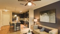This single-story space showcases an open-concept design that seamlessly connects the kitchen, dining, and living areas into one cohesive and functional layout. The kitchen is outfitted with dark espresso cabinetry that extends to the upper walls, stainless steel appliances including a refrigerator, range, and built-in microwave, and a granite-topped island with bar seating — ideal for casual dining or entertaining. Track lighting above the kitchen provides focused task illumination, while pendant lights suspended over the island add a decorative touch.

The living area transitions naturally from the kitchen and dining space, anchored by a ceiling fan with an integrated light fixture and defined by a bold, dark accent wall that adds depth and contrast. Crown molding lines the ceiling, contributing a refined architectural detail throughout. Warm-toned flooring runs across the shared space, enhancing the inviting ambiance. The living room is well-proportioned, offering comfortable furniture arrangements beneath soft ambient lighting. Together, the spaces create a warm, contemporary atmosphere suited to both relaxed everyday living and social gatherings.