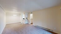 This single-floor residential unit offers a practical and comfortable layout designed for everyday living. The open living and dining area is generously proportioned, featuring neutral-toned carpet, warm beige walls, and a baseboard heating system. A chandelier accents the dining area, which flows seamlessly into the kitchen via a peninsula breakfast bar. The kitchen is well-equipped with white shaker-style cabinetry, granite-look countertops, stainless steel appliances including a full-size refrigerator, and light wood-look flooring that extends into an adjacent laundry nook housing a washer and dryer. The bedroom is a comfortable retreat with plush carpeting, clean neutral walls, and a large double-door closet providing ample storage. An en-suite bathroom features white cabinetry with granite countertops, a vanity mirror, and a full bathtub and shower combo with tile surround. The unit also includes a dedicated bathroom accessible from the main living space, maintaining a similar clean, white aesthetic. Throughout, the space is characterized by its clean lines, neutral color palette, and functional design, making it a move-in-ready home with thoughtful amenities.
