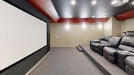 Located at 739 S 300 W in Salt Lake City, Utah, this purpose-built home theater room delivers a true cinematic experience within a compact, efficiently designed space. The room is defined by its bold design choices: a dark, textured ceiling punctuated by recessed spotlights creates an immersive atmosphere, while a striking deep red accent band wraps the upper perimeter of the walls, adding a dramatic, theater-inspired aesthetic. A large projection screen is mounted flush against one wall, flanked by a black surround that enhances contrast and focus during viewing. Ceiling-mounted projector infrastructure is in place, ensuring a clean, professional setup. The seating area features a raised platform with two rows of plush, dark leather reclining chairs, providing excellent sightlines from every seat. Wall-mounted sconce lighting adds a warm, ambient glow along the neutral-toned side walls, while multiple in-wall speaker cutouts indicate a comprehensive surround sound system. Carpet flooring throughout contributes to both comfort underfoot and acoustic dampening. The overall design is cohesive and purpose-driven, making this space a well-appointed private screening room suited for an engaging home entertainment experience.