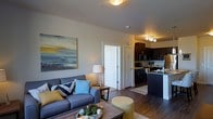 This single-floor residence showcases an open-concept layout that seamlessly connects the living area and kitchen, creating a spacious and functional environment. The living room is anchored by a comfortable grey sofa accented with blue and yellow decorative pillows, complemented by a rustic wood coffee table and a warm table lamp that adds ambient lighting. A large abstract canvas painting in yellow and grey tones adorns the wall, adding a contemporary artistic touch to the space.

The kitchen is equipped with dark espresso cabinetry that contrasts cleanly against lighter countertops, stainless steel appliances, and a peninsula-style island that doubles as a breakfast bar. A track lighting fixture overhead provides ample task lighting throughout the kitchen area. Rich hardwood-style flooring flows continuously through both the living and kitchen spaces, enhancing the cohesive feel of the open floor plan.

A doorway visible from the living area leads to additional rooms, suggesting a practical and well-organized layout. The neutral warm-beige wall tones throughout contribute to a welcoming and modern ambiance, making the space feel bright, livable, and well-designed.