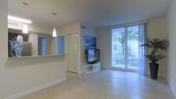 This single-floor space is designed with a clean, contemporary aesthetic that emphasizes openness and natural light. The living area serves as the heart of the unit, anchored by large floor-to-ceiling sliding glass doors that lead to a private balcony. Lush tropical foliage is visible beyond the balcony railing, creating a serene, resort-like atmosphere. Light-toned tile flooring runs throughout, reflecting the abundant natural light and enhancing the airy feel of the space. A wall-mounted flat-screen television is positioned on a sleek, curved media console, adding a modern touch to the living area. Adjacent to the main living space, a partial wall with a pass-through counter connects to what appears to be a kitchen area, where a pendant light fixture hangs above the countertop, complementing the neutral, cream-toned color palette. Built-in storage with louvered doors provides practical organization while blending seamlessly with the surrounding walls. Overall, the space offers a bright, well-organized layout with a warm tropical ambiance.