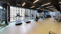 This single-floor fitness center is thoughtfully designed to accommodate a full range of exercise disciplines within a spacious, open layout. The facility is divided into distinct functional zones, each purpose-built for specific training needs. The cardio area is lined with a bank of floor-to-ceiling windows overlooking the surrounding cityscape, housing a row of treadmills, ellipticals, and stationary bikes — all from Life Fitness — allowing users to exercise with natural light and urban views. Adjacent to this is a comprehensive strength training zone equipped with free weights, dumbbells, adjustable benches, and a Smith machine, also benefiting from large windows that open to outdoor greenery. A dedicated functional training studio provides an open, flexible space outfitted with stability balls on a multi-tier rack, a BOSU trainer, and step platforms, ideal for group classes or individual functional workouts. Throughout the facility, durable resilient flooring in neutral tones provides a clean, practical surface, while the dark suspended ceiling panels and linear LED lighting lend a modern, polished aesthetic. The facility also includes a water fountain and direct access to an outdoor area, enhancing the overall workout experience.