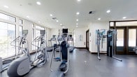 This fitness center offers a thoughtfully designed workout space spread across a single level. The room is outfitted with a selection of cardio equipment, including elliptical trainers, treadmills, and stair climbers, all arranged to allow comfortable movement between machines. The flooring transitions between a durable dark gym surface in the main exercise area and a lighter finish near the entry, providing functional zoning within the space.

Large windows line the walls, allowing generous natural light to fill the room while offering views of the surrounding greenery outside. Recessed lighting installed throughout the ceiling ensures the space remains well-lit regardless of the time of day. The walls are painted a clean, neutral white and feature decorative artwork, contributing to a welcoming atmosphere that balances utility with aesthetics.

A fire safety station is visible along one wall, reflecting attention to safety standards. The overall ambiance is bright, open, and organized, making it a practical and inviting environment for fitness activities.