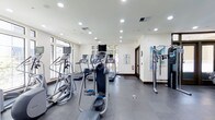 This fitness center offers a thoughtfully designed workout space spread across a single level. The room is outfitted with a selection of cardio equipment, including elliptical trainers, treadmills, and stair climbers, all arranged to allow comfortable movement between machines. The flooring transitions between a durable dark gym surface in the main exercise area and a lighter finish near the entry, providing functional zoning within the space.

Large windows line the walls, allowing generous natural light to fill the room while offering views of the surrounding greenery outside. Recessed lighting installed throughout the ceiling ensures the space remains well-lit regardless of the time of day. The walls are painted a clean, neutral white and feature decorative artwork, contributing to a welcoming atmosphere that balances utility with aesthetics.

A fire safety station is visible along one wall, reflecting attention to safety standards. The overall ambiance is bright, open, and organized, making it a practical and inviting environment for fitness activities.