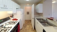 This single-story space is designed with a practical, functional layout that makes efficient use of its footprint. The floor plan, visible in the 3D model, reveals a thoughtfully arranged interior that includes a living area, kitchen, bedroom spaces, and a bathroom.

The carpeted rooms are finished in soft, neutral beige tones with clean white baseboards, creating a calm and cohesive atmosphere. One room features a window fitted with horizontal blinds, allowing natural light to filter in while maintaining privacy. A second carpeted room includes two doors—one leading to what appears to be a walk-in closet or storage area, and another providing additional access, offering a well-organized layout suitable for a bedroom.

The galley-style kitchen is outfitted with white cabinetry, white appliances including a gas range and refrigerator, a double stainless steel sink, and durable tile flooring in an earthy stone pattern. A utility access panel is visible in the adjoining hallway, adding practical functionality.

The bathroom includes a single-sink vanity with white cabinetry, a large mirror with overhead lighting, a shower-tub combination, and tile flooring, completing the space with clean, straightforward finishes throughout.