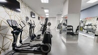Located at 145 Granby St in Norfolk, Virginia, this fitness center offers a thoughtfully designed workout environment within a compact yet efficiently utilized single-floor layout. The space is defined by its striking geometric accent wall — a bold white and gold angular pattern that adds a modern, upscale aesthetic throughout the room. Recessed flush-panel ceiling lights provide bright, even illumination across the polished concrete floors, creating a clean and energizing atmosphere.

The cardio section is well-appointed with multiple elliptical machines and treadmills, including Precor and SportsArt models, each equipped with individual monitors for entertainment during workouts. A Smith machine and free weight area with a tiered dumbbell rack and adjustable bench serve the strength training needs of users. A cable resistance station and additional weight equipment round out the functional fitness offerings.

A floor-to-ceiling mirror spans one wall, visually expanding the space and allowing users to monitor their form during exercise. Multiple wall-mounted flat-screen televisions are positioned throughout for easy viewing from all equipment stations. The space also features a dedicated free weights zone with medicine balls and a workout bench. Clean white walls, minimal decor, and organized equipment placement contribute to an uncluttered, professional-grade fitness environment.