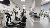 Located at 145 Granby St in Norfolk, Virginia, this fitness center offers a thoughtfully designed workout environment within a compact yet efficiently utilized single-floor layout. The space is defined by its striking geometric accent wall — a bold white and gold angular pattern that adds a modern, upscale aesthetic throughout the room. Recessed flush-panel ceiling lights provide bright, even illumination across the polished concrete floors, creating a clean and energizing atmosphere.

The cardio section is well-appointed with multiple elliptical machines and treadmills, including Precor and SportsArt models, each equipped with individual monitors for entertainment during workouts. A Smith machine and free weight area with a tiered dumbbell rack and adjustable bench serve the strength training needs of users. A cable resistance station and additional weight equipment round out the functional fitness offerings.

A floor-to-ceiling mirror spans one wall, visually expanding the space and allowing users to monitor their form during exercise. Multiple wall-mounted flat-screen televisions are positioned throughout for easy viewing from all equipment stations. The space also features a dedicated free weights zone with medicine balls and a workout bench. Clean white walls, minimal decor, and organized equipment placement contribute to an uncluttered, professional-grade fitness environment.
