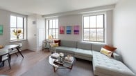 Located on Broad Street in the heart of Newark, NJ, this two-bedroom, one-bathroom residence offers a well-designed layout with modern finishes throughout. The open living area is bright and airy, featuring hardwood flooring, neutral walls, and large grid-pane windows that frame impressive panoramic city views. A sectional seating area anchors the living room, which flows naturally into the contemporary kitchen. The kitchen is outfitted with light wood cabinetry, white quartz countertops, stainless steel appliances including a full-size refrigerator, electric range, and microwave, along with a geometric white tile backsplash and recessed lighting. The primary bedroom features dual windows with city views, hardwood floors, and a calm gray color palette. The second bedroom offers a bold blue accent wall complemented by decorative wall paneling, warm wood nightstands, and a window with natural light. The bathroom is finished with herringbone-patterned tile flooring, a floating vanity with a large mirror, and clean white walls, creating a crisp, modern aesthetic. Throughout the residence, the design balances contemporary style with functional living in an urban setting.