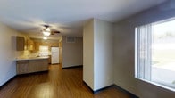This single-floor residential unit offers a practical and comfortable layout designed for everyday living. The space features warm-toned hardwood flooring throughout, adding a sense of continuity and warmth across the open living area and connecting hallway.

The kitchen is equipped with honey-toned wood cabinetry, a white refrigerator, and a range, providing a functional and classic cooking environment. A ceiling fan with an integrated light fixture serves the kitchen and adjacent living area, ensuring good air circulation and ambient lighting. The textured popcorn ceiling adds a traditional finish to the interior.

A central hallway connects the main living space to the rest of the unit, flanked by a structural wall that helps define the open living area. Neutral beige wall tones throughout create a clean, understated backdrop that suits a variety of décor styles. A thermostat panel and HVAC vent are conveniently positioned along the hallway wall, reflecting thoughtful placement of functional elements.

Natural light filters in through windows on the side of the living area, brightening the space and complementing the warm flooring tones. The overall layout is straightforward and efficient, making it well-suited for comfortable day-to-day living.
